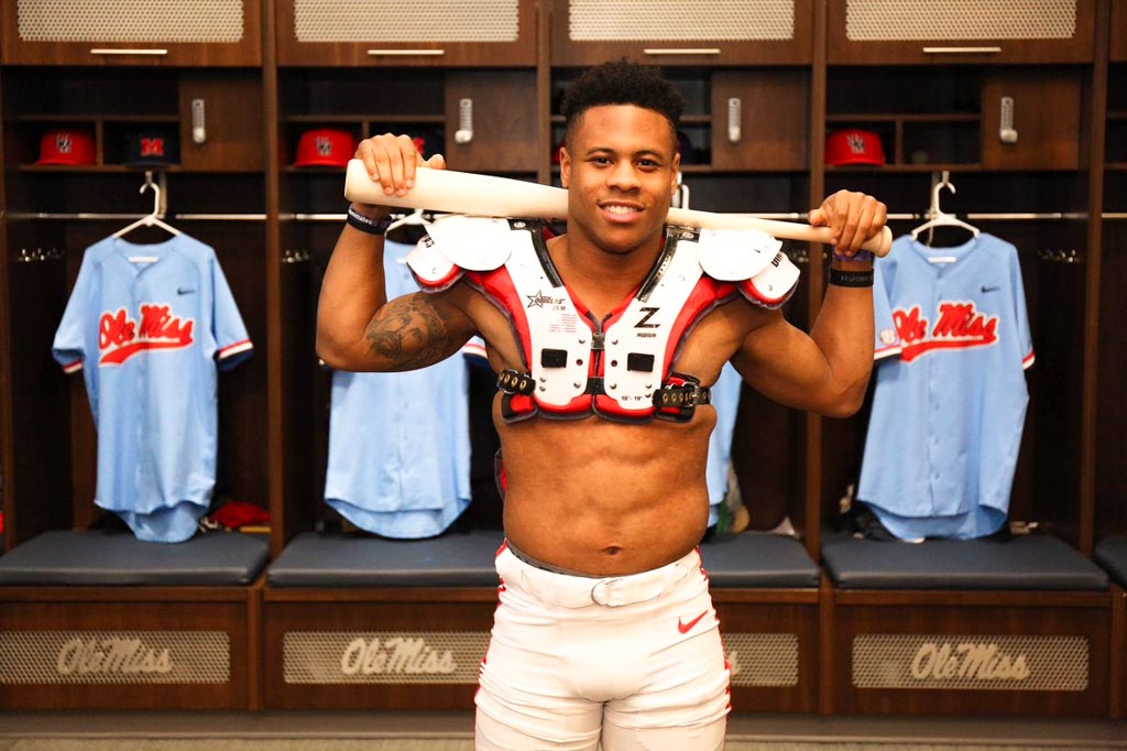 Ole Miss running back Jerrion Ealy poses in locker room wearing shoulder pads and holding a baseball bat over his shoulders.